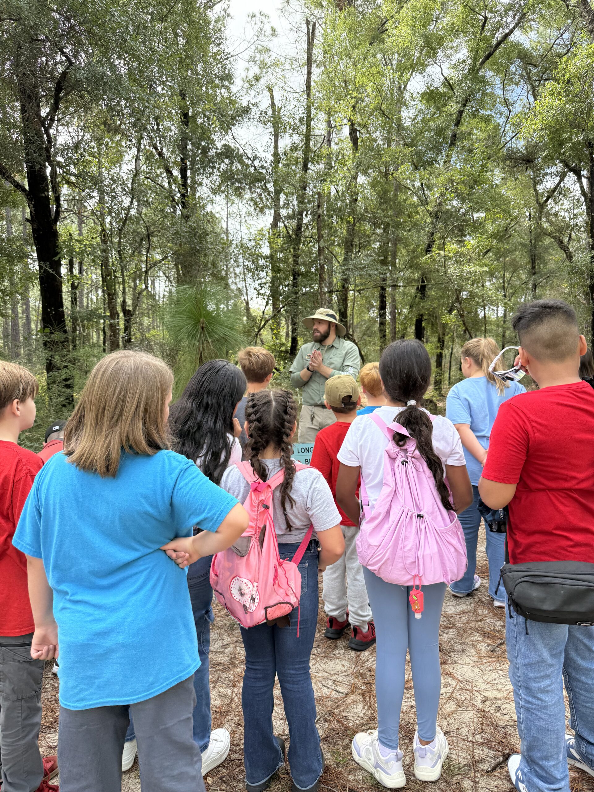 Group of kids with colorful backpacks listening to a bearded man in a wide-brimmed hat giving an outdoor talk in a forest clearing.
