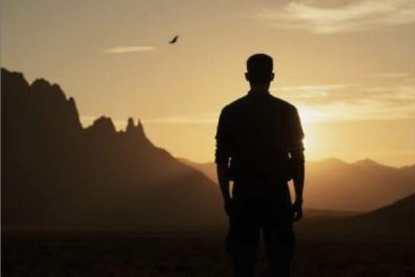 Silhouette of a man standing in a desert at sunset with rocky mountains in the distance.
