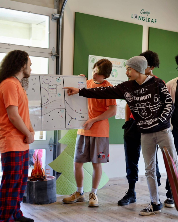 Four young people in orange shirts stand around a whiteboard with diagrams, one pointing at it during a camp activity at Camp Longleaf.