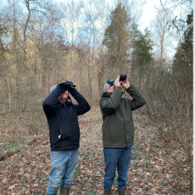 Two people standing side by side in a leaf-covered forest, wearing jackets and boots, looking up through binoculars at something in the trees.