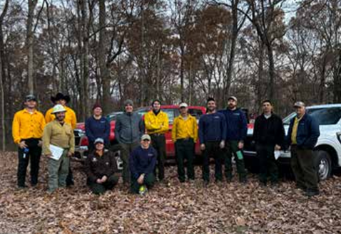 Group of volunteers posing in a leaf-strewn forest clearing, many wearing yellow jackets, with vehicles in the background.