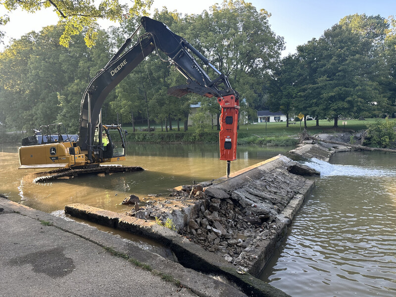 Yellow excavator with hydraulic hammer breaking a concrete barrier in a pond near a grassy, tree-lined bank.