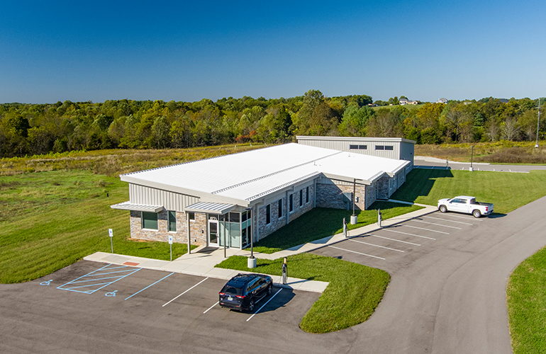 A single-story stone-and-metal building with a white metal roof, surrounded by green fields and a clear blue sky, with visible parking lot and handicapped spots in front.