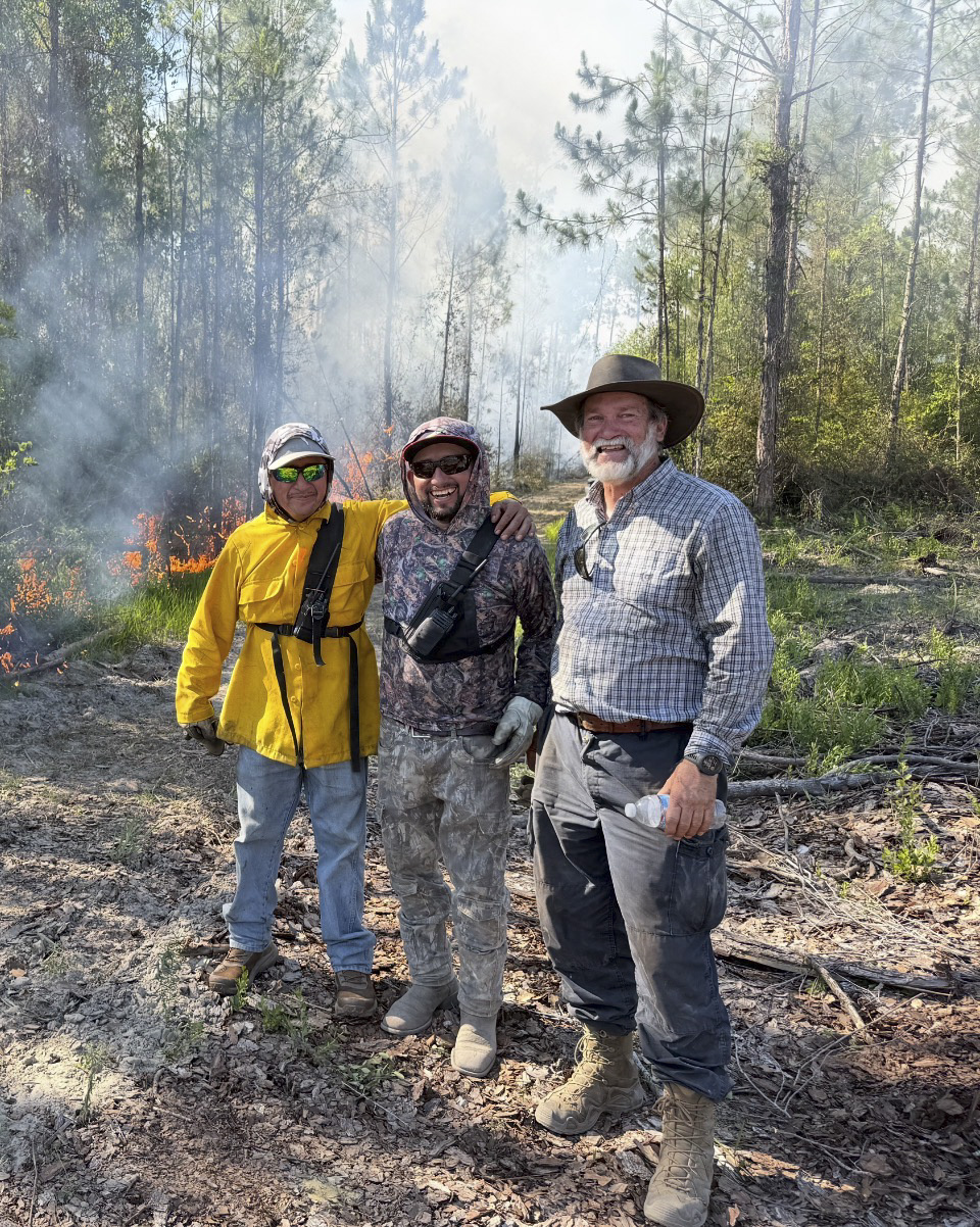 Three men stand together in a smoky forest fire area, two in protective gear and hats smiling at the camera.