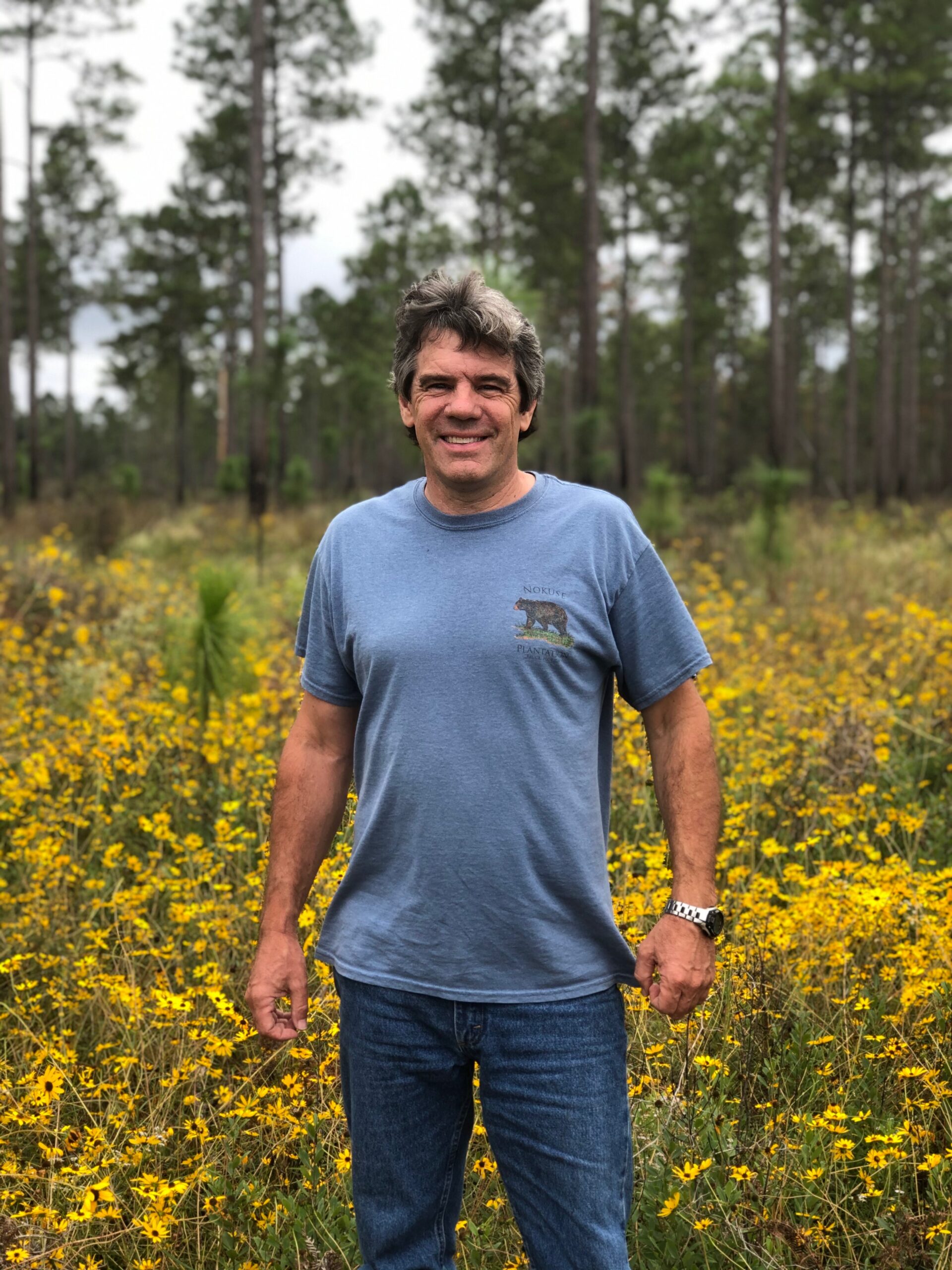 Man in a blue t-shirt standing in a field of yellow wildflowers with tall trees in the background, smiling at the camera.