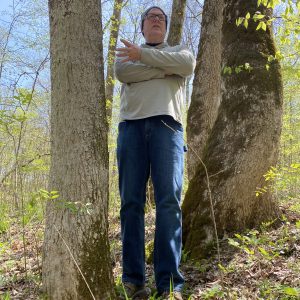 Person wearing a beanie and glasses stands between two large trees in a sunlit forest, arms crossed.