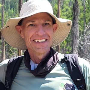 Person wearing a beige wide-brimmed sun hat and a hiking backpack, smiling outdoors in a forested area.