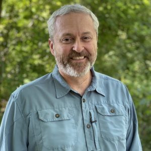 Smiling man with gray hair and beard outdoors, wearing a teal button-up shirt.